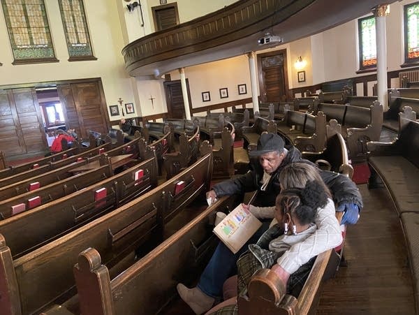 Two adults and one child sit in a church pew while looking at a picture book. 