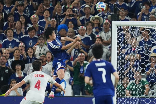 A soccer player in blue jumps for a header, with the crowd in the background.