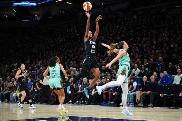 A basketball player in black leaps for a jump shot between two players in turquoise.