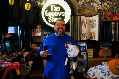 A man in a Chicago Cubs shirt holds a dog while smiling for a photo in his shop