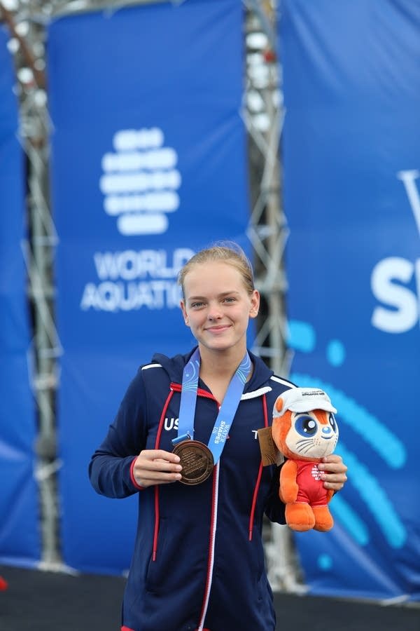 A diver shows off her bronze medal while smiling and holding an orange plush doll.