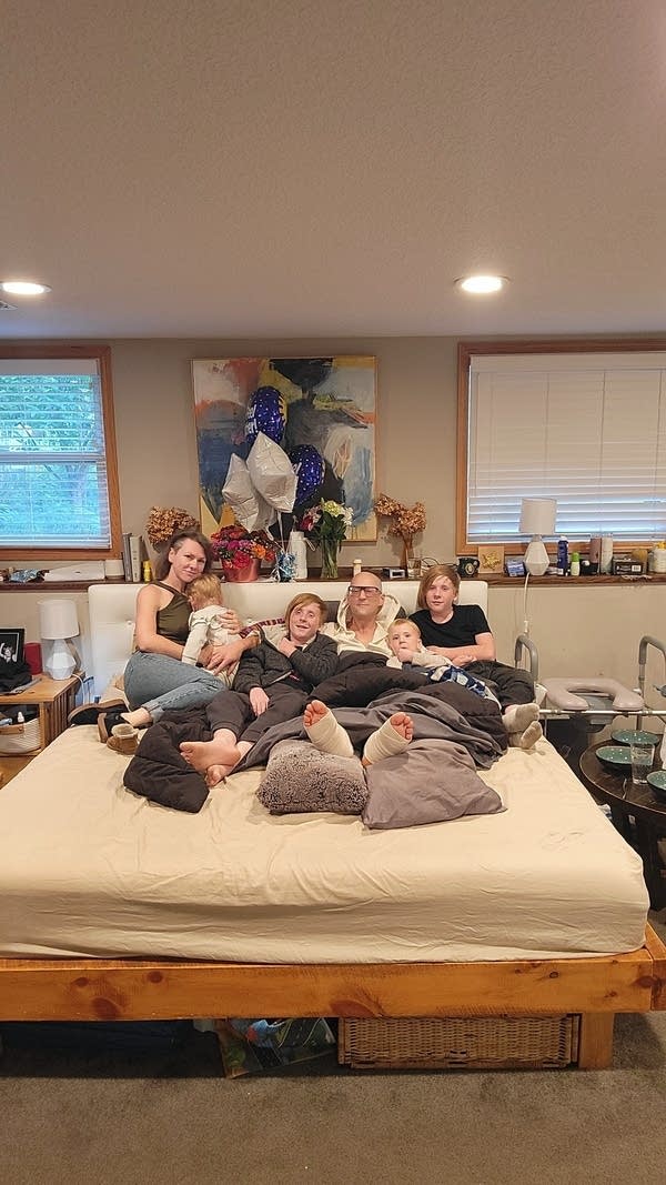 A family poses for a photo in a bed. The man in the center has casts around both feet while he recovers from injuries.