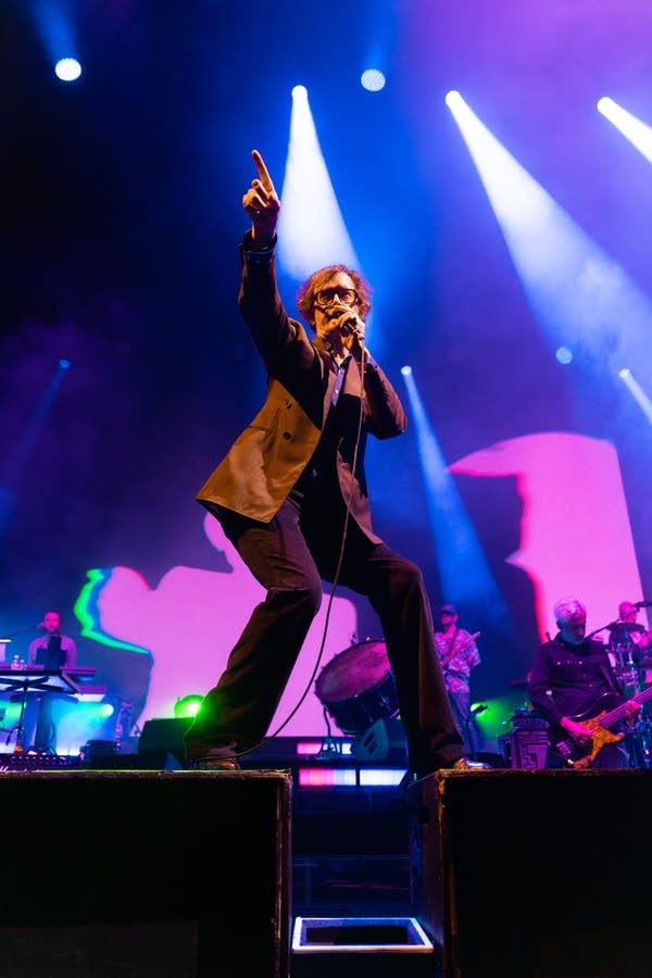 Jarvis Cocker of Pulp, impeccably dressed in a brown suit, points upward as he sings into a microphone at Armory.