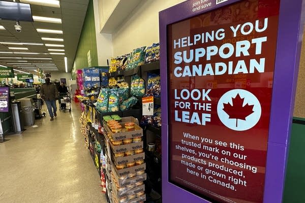 A red digital sign in a grocery store instructs shoppers to "look for the leaf" when buying goods to support Canadian businesses and avoid paying for imports with tariffs. The shelf next to the sign has chips and snacks.