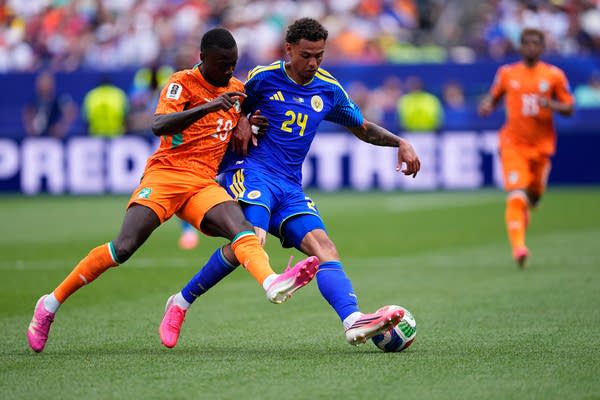 A soccer player in orange and a player in blue fight for the ball.