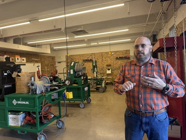 A bald man with a beard wears blue jeans with an orange and blue button-up speaks in a workshop-like setting.
