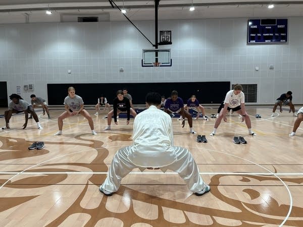 A man in long white pants and a shirt, his back to the camera, squats with legs wide apart on a gym floor. Several young adults face him while imitating the pose. (Jennifer Pak/Marketplace)