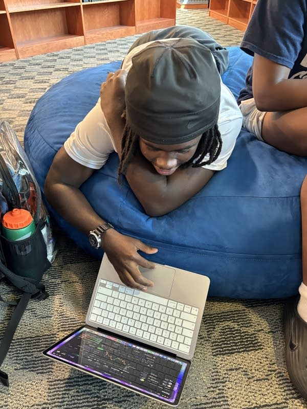 A high schooler in a grey cap checks stocks on an open laptop. 