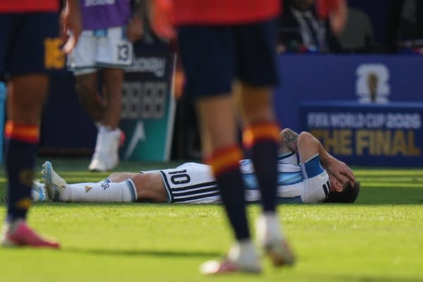A soccer player lies on the ground, covering his face with his hands, as other players look on.