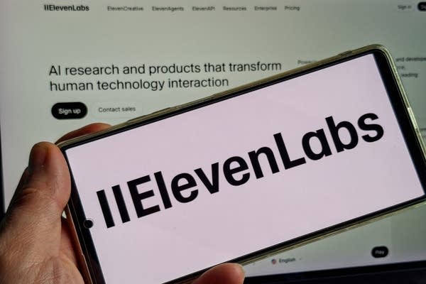 ElevenLabs' logo displayed on a phone screen with a laptop behind