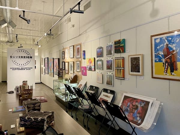Wide shot of a gallery. Some works hang on walls while others are stacked on stands.