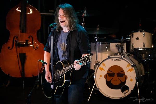 Dave Pirner plays a black acoustic guitar while singing into mic
