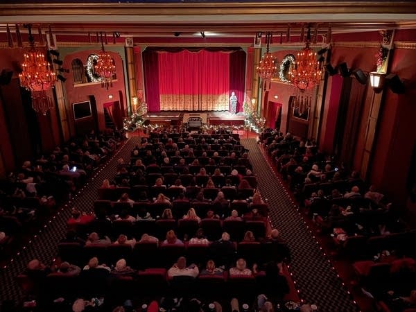 Wide aerial shot of a packed theater watching a man on stage.