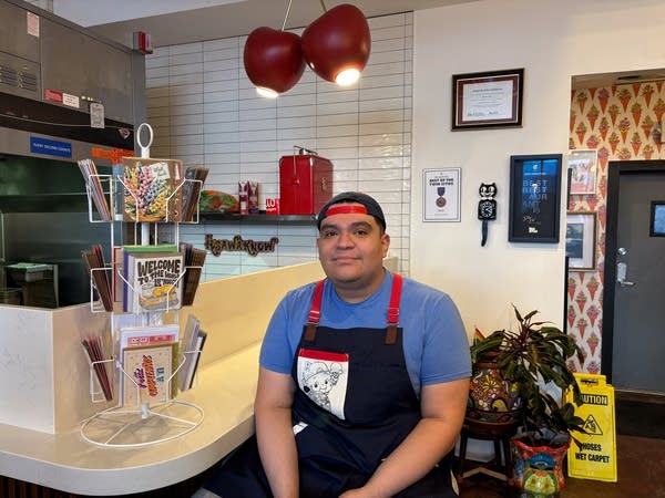A man in an apron and baseball cap poses in an ice cream shop.