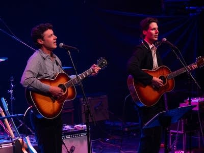 Two men sing into microphones and play acoustic guitars.