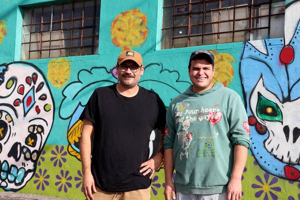 Two men pose for a photo in front of a brightly colored mural on a warehouse wall.