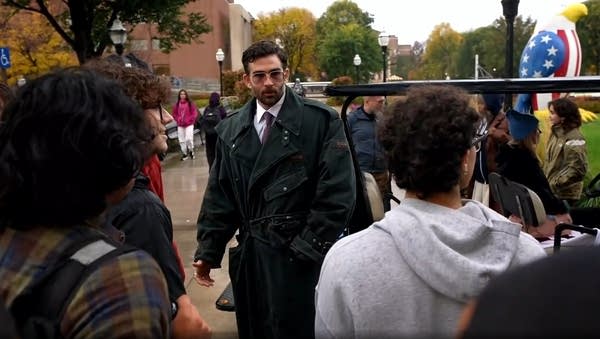A man in a dark green trench coat addresses a crowd of college students.