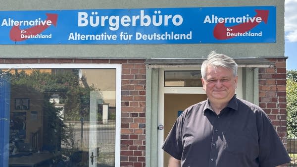 A man smiles in front of a sign for AfD