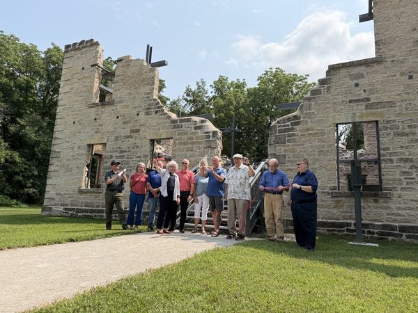 A group of people cheer after a ribbon cutting next to ruins of a limestone building.