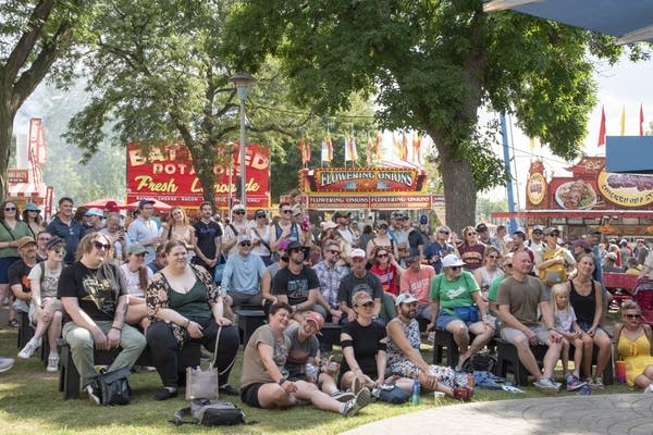 A crowd of people fill a seating area near an outdoor stage at a fair