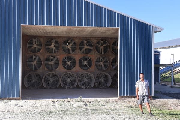 Wide shot of a man posing for a photo in front of a blue barn. Rows of fans are seen inside.