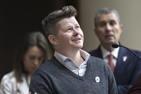 Becca Good at a news conference wearing a sweater and collared shirt