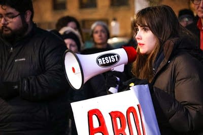 A woman holds a protest sign and speaks into a bullhorn.