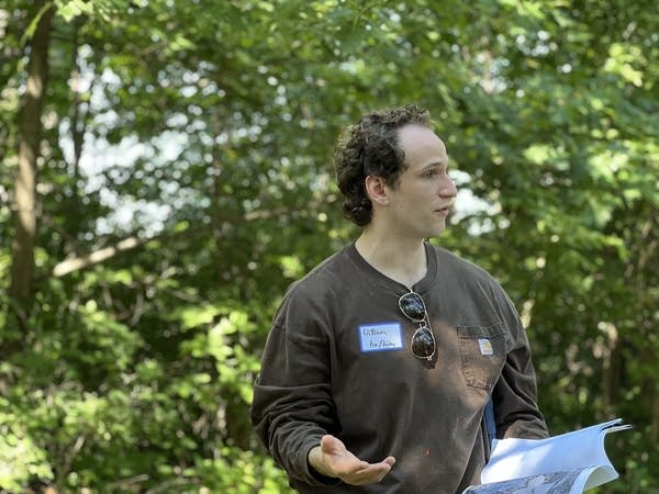A man speaks to a group while standing in front of a tree line.