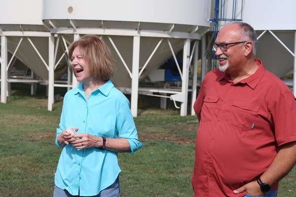 A woman in a light blue shirt and a man in a red shirt stand together and laugh on a farm.
