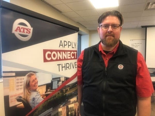 A man stands next to a company sign that reads "Apply Connect Thrive."