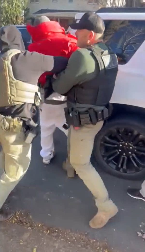 Two men in tactical gear grab a man in red and pull his hands behind his back.
