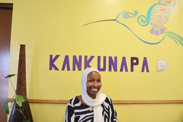 A woman smiles next to a yellow wall with "Kankunapa" painted in purple and an illustration of a hummingbird.