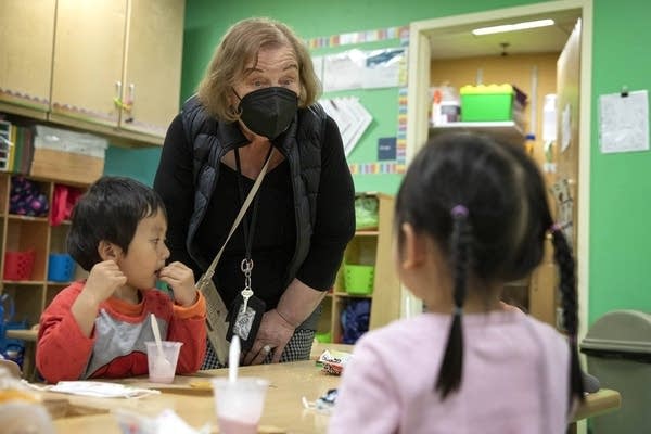 A woman with a black shirt, black puff vest and black face mask looks expressively at two young children sitting at a wood table, eating.