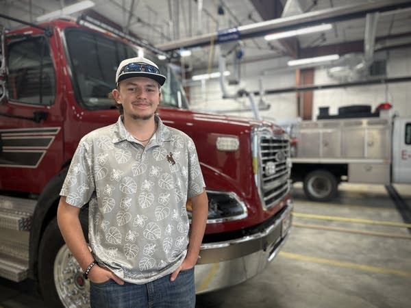 A teenager in a grey and white polo shirt and ball cap. He's standing in front of a red fire truck.