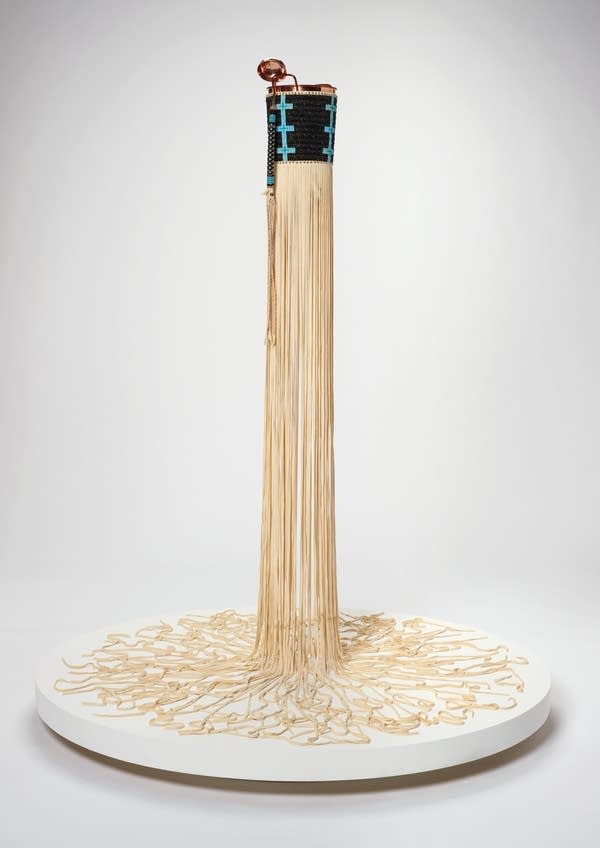 A sculpture of beige strands coming down from a black and teal cyllinder.