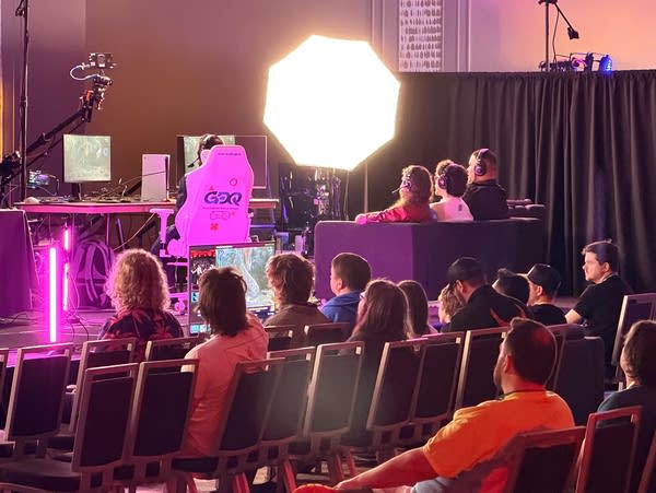 An audience watches a man in a white gaming chair play a video game with a couch of three commentators behind him.