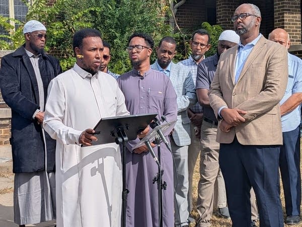 A man speaks at a lectern in front of a group of people outside a mosque.