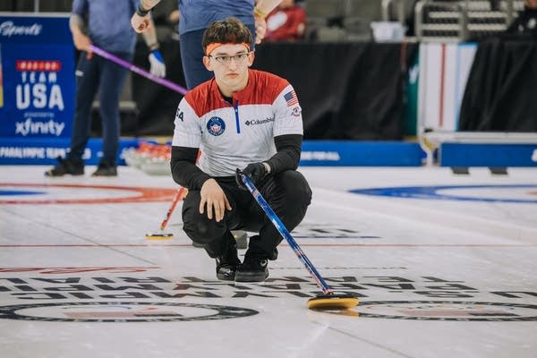 A curler kneels on the ice to watch his throw.