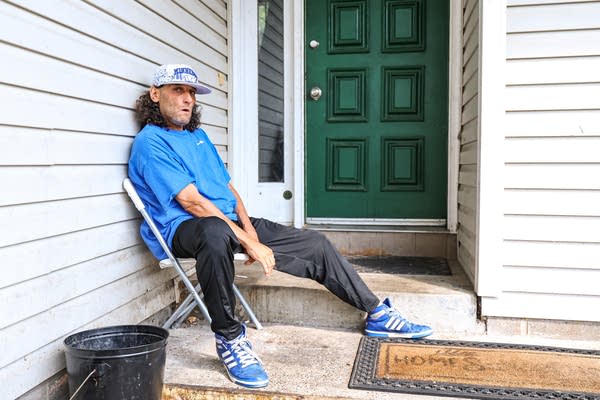 A man in a blue shirt sits in a folding chair on the front porch.