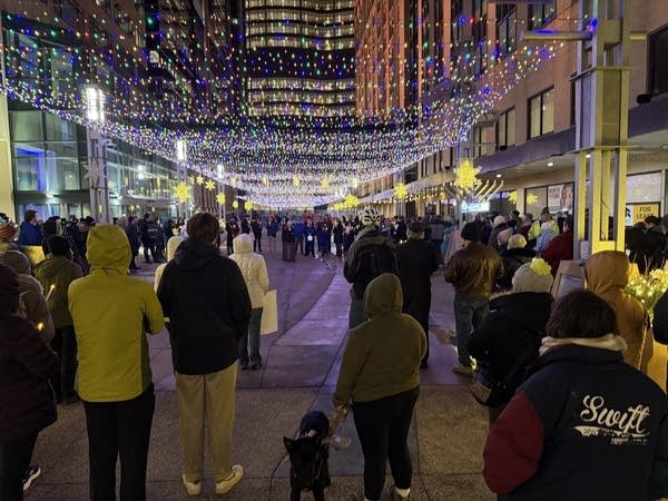 A crowd gathers for a vigil in a downtown plaza under a canopy of Christmas lights.