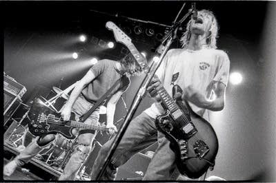 Kurt Cobain sings into a microphone and strums a guitar.