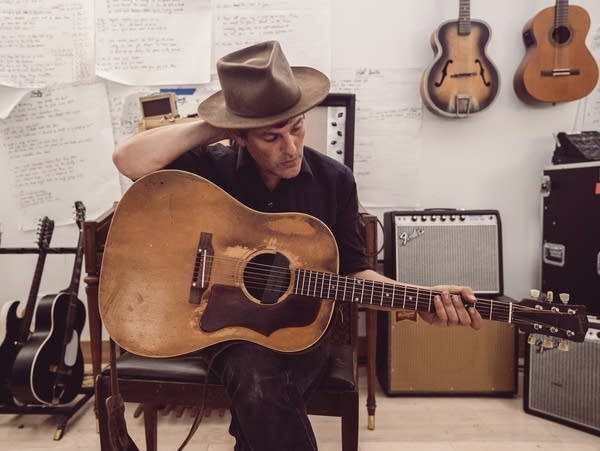 A man sits on a chair holding an acoustic guitar.