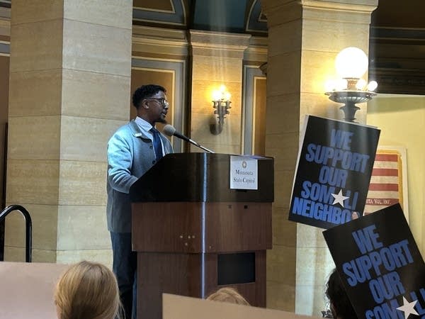 A man speaks at a podium in front of signs reading "We support our Somali neighbor."