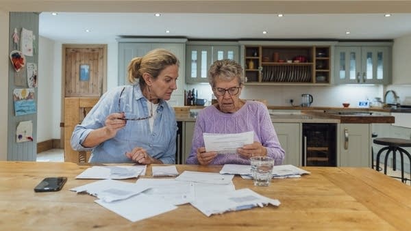 A senior woman and her mature daughter at home in Seghill, Northumberland. The senior woman is staying with her daughter for support and care as she has dementia. They are sat at the dining room table where they're looking through financial bills, looking concerned.