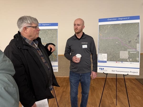A man listens while standing next to maps on display. He's holding a coffee cup.