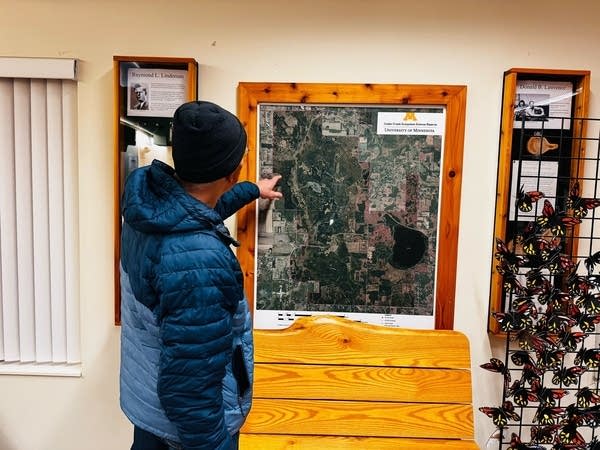 A man in a coat and hat points to a framed map on the wall.