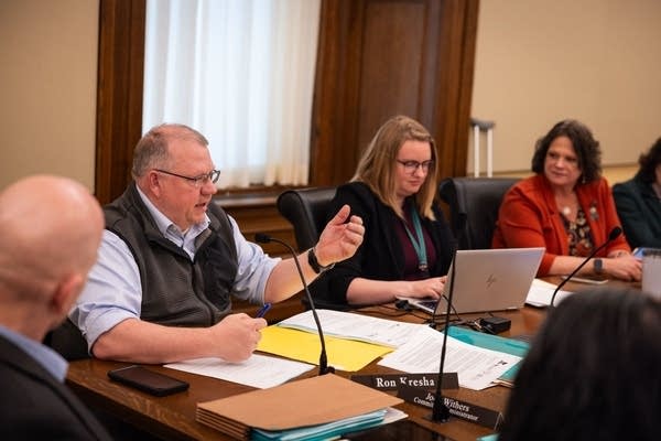 A group of people sit at a table with microphones and documents. One speaks with his hand held up.