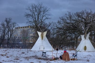 A woman sits near a fire, two tipis are in the background.