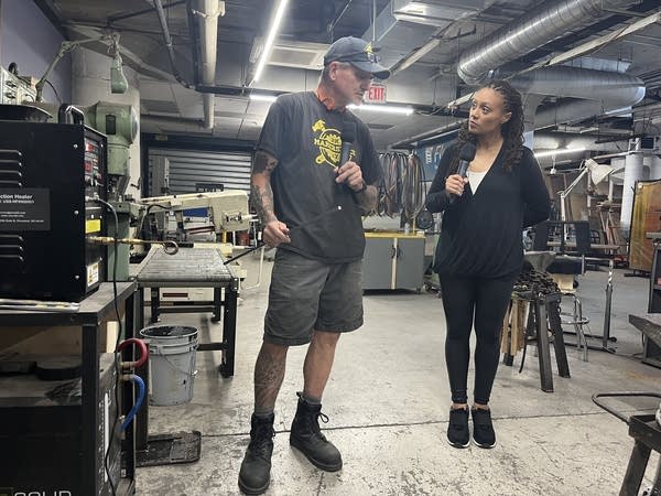 Scott Van Campen (left), executive director of MakerSpace NYC, holds up a metal rod for inspection by Kimberly Adams, "Marketplace Morning Report" host. They're standing next to an induction forge.