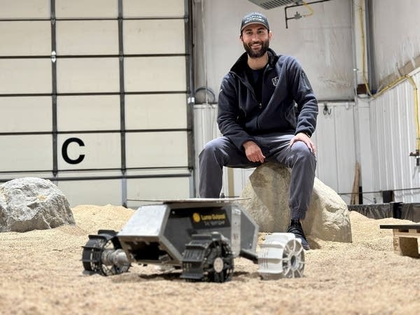A man with a dark beard, dressed in a blue jacket and baseball hat, sits on a rock in a sandy area inside a warehouse. In the foreground is a small silver and gray rover with four wheels.
